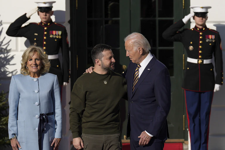 Two men face towards each other with their arms around each other. One is older and has on a blue suit. The other has short brown hair and green camo shirt. They stand in front of a woman with. blue suit and two soldiers behind them saluting.