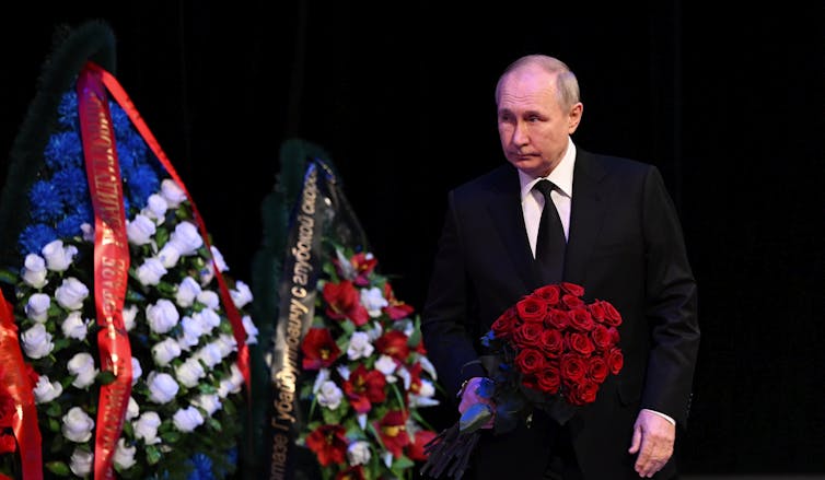 A balding man walks onto a stage festooned with flower arrangements.