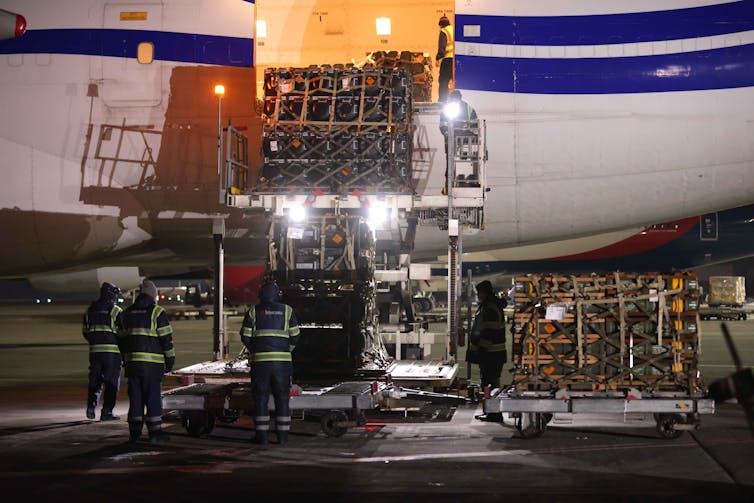 People with reflective vests and dark clothing are seen unloading large boxes from an airplane at nighttime.