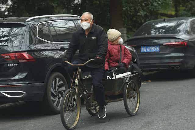 An elderly Chinese man pedals a tricycle with an elderly women in the back.