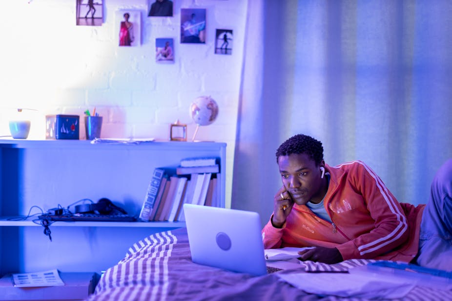 A young man in a red zip-up sweatshirt and casual pants lies on a bed looking at a laptop