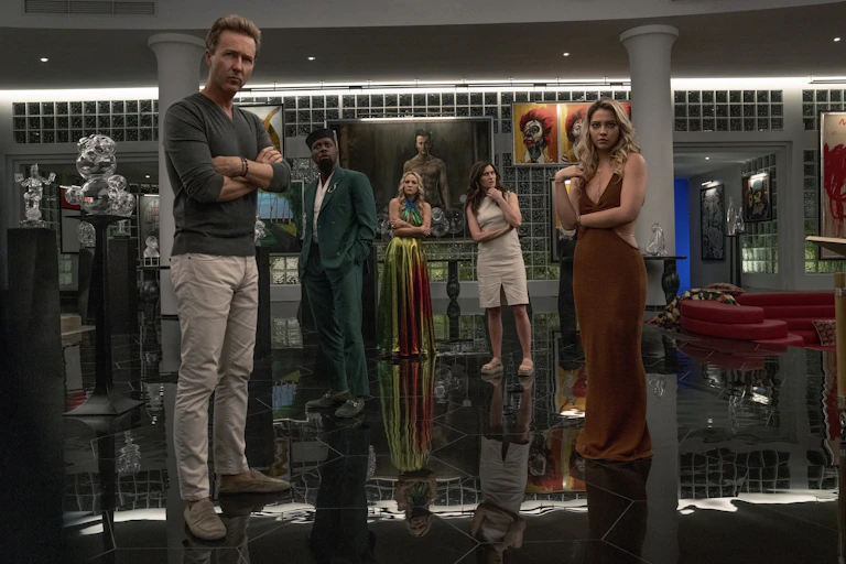 Miles Brown and his friend in Knives Out, all standing dramatically with arms folded in a lobby filled with art and glass sculptures