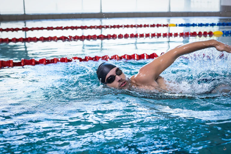 A man wearing a swimming cap and goggles swims in an indoor pool.