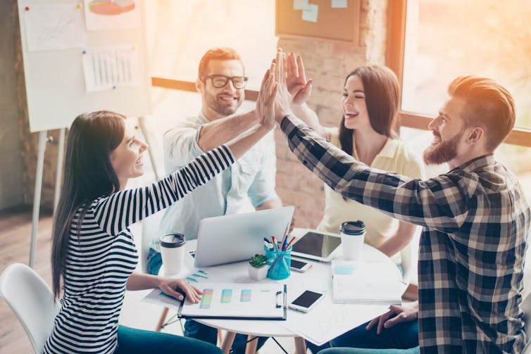 In a sunny office, four young people smile and give each other high fives around a table of laptops and charts