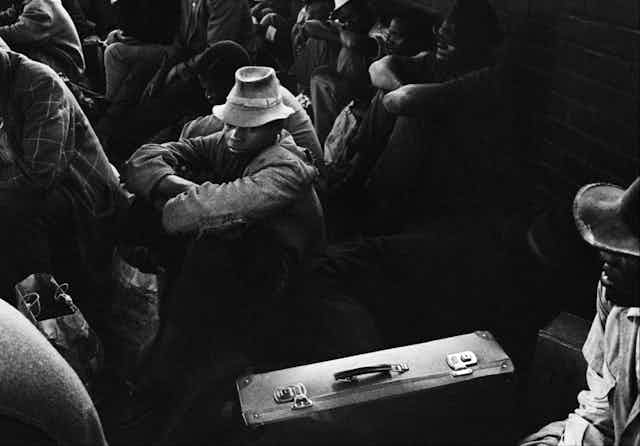 A moody black and white photo of a group of men sitting. The focus is on a young man in a hat, his suitcase next to him, looking grim.