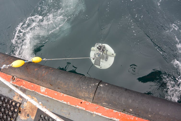 A drum covered in electronic devices is lowered over the side of a boat into the ocean.