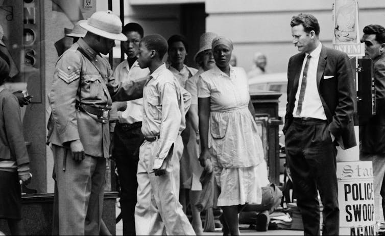 A policeman in old-fashioned uniform puts a hand on the shoulder or a young man in worker's clothes. Men in suits look on idly, hands in their pockets.