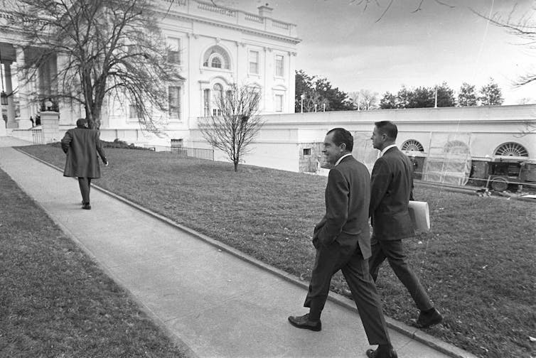 Two men in suits walking on a path toward the White House.