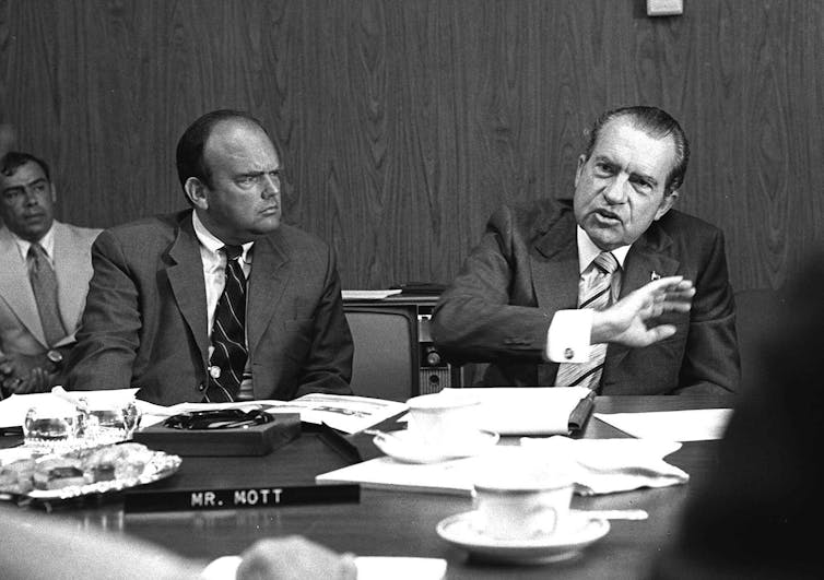 Two men in suits sitting at a table strewn with papers.