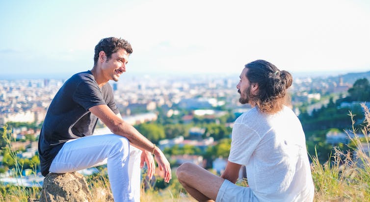 Two men sit on a green hill talking with a city below them
