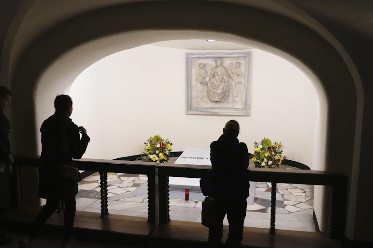 The silhouette of two people standing before a tomb which has flowers on it and a painting above it.