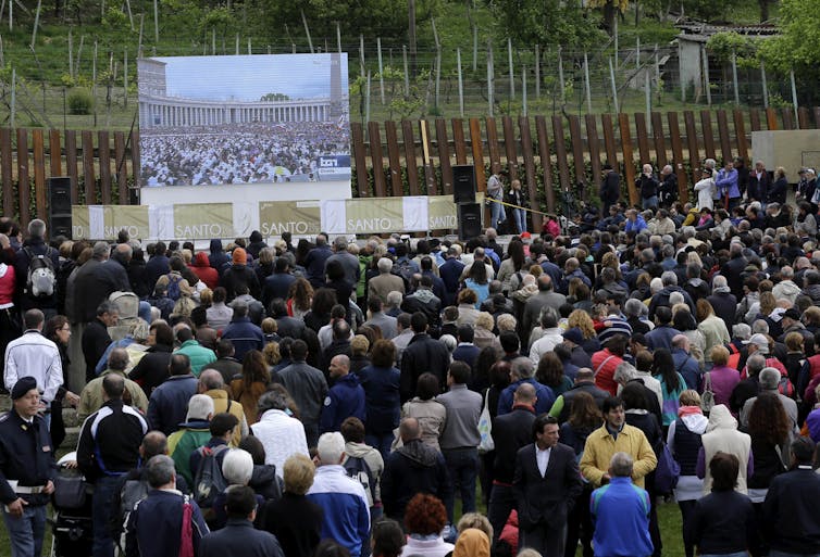 Hundreds of people gathered outside watching a ceremony on a large screen.