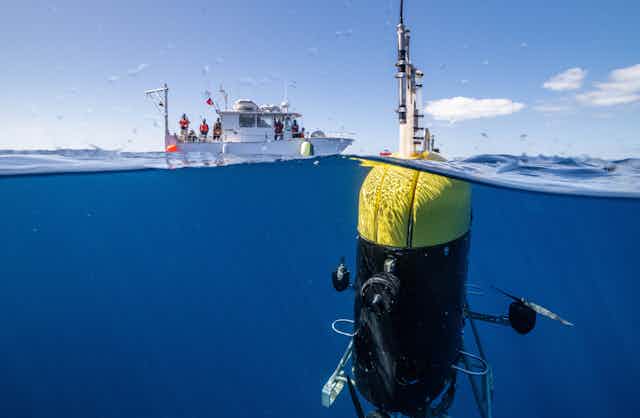 A large robot loaded with sensors is shown with its antenna above water and the rest beneath the surface, with a research ship in the background.