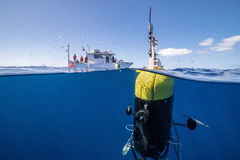 A large robot loaded with sensors is shown with its antenna above water and the rest beneath the surface, with a research ship in the background.