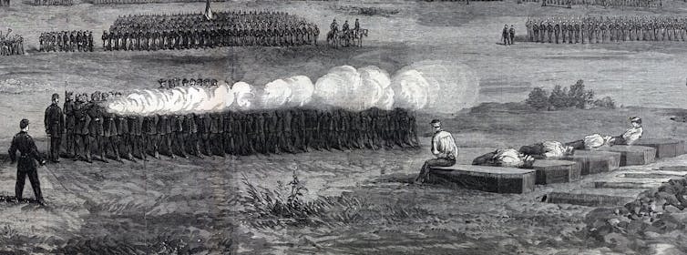A large firing squad executing five men sitting at the foot of their coffins.