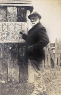 A black and white image of a First Nations man, William Barak stands before a painting. He is wearing a hat and dark jacket.