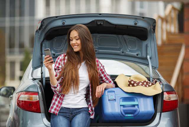 A young woman smiles at her mobile phone while resting on the open trunk of a car, filled with a suitcase and hat