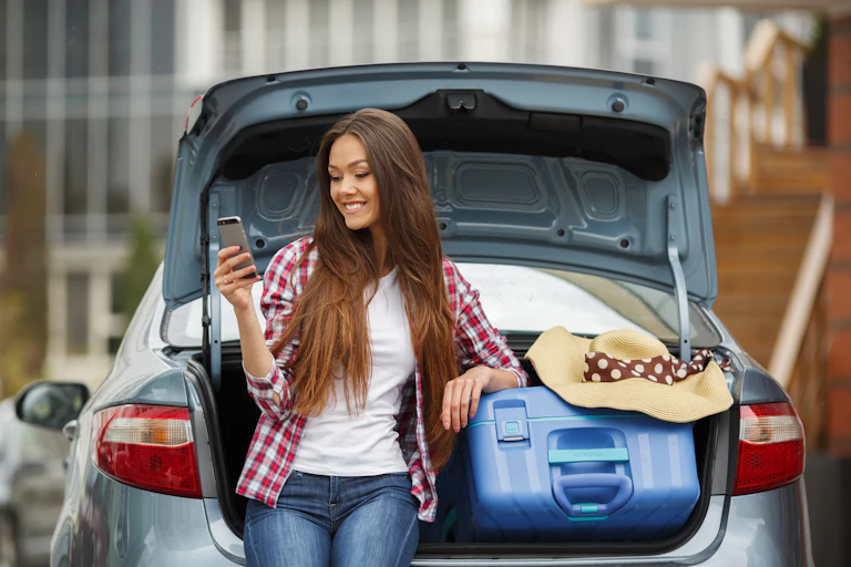 A young woman smiles at her mobile phone while resting on the open trunk of a car, filled with a suitcase and hat