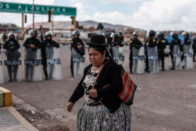 An indigenous Peruvian woman walks across a road in front of a row of police with riot shields