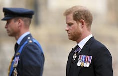 A man in a military uniform and hat stands blurred in the background while a red-haired man with a beard walks beside him in the foreground.