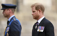 A man in a military uniform and hat stands blurred in the background while a red-haired man with a beard walks beside him in the foreground.