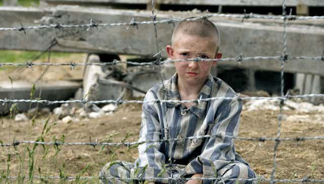 A boy sits cross legged behind barbed wire wearing blue and white striped pyjamas.