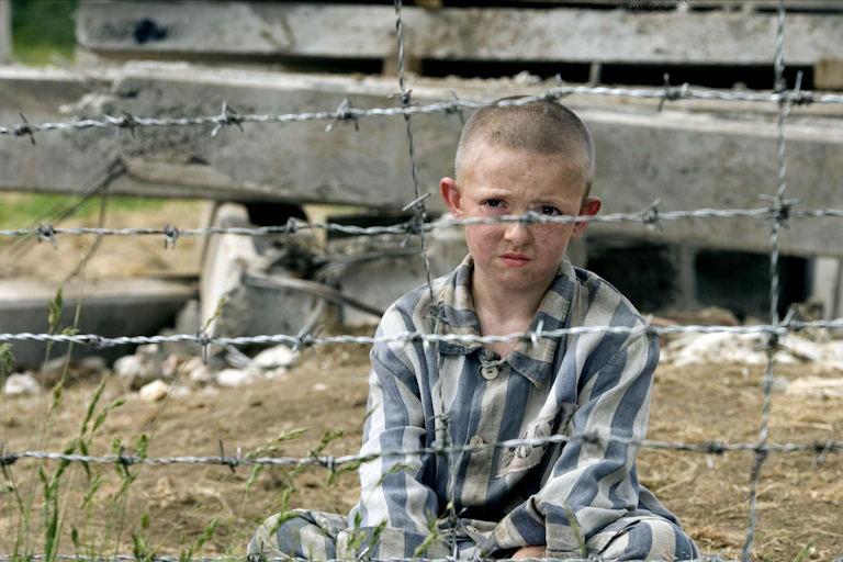 A boy sits cross legged behind barbed wire wearing blue and white striped pyjamas.