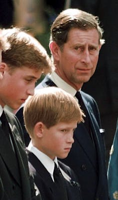 A dark haired man frowns standing next to two boys, one a teenager looking down and a smaller boy with red hair staring ahead.