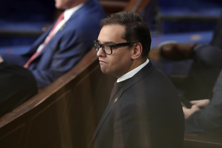 Close up of a young man wearing black rimmed glasses and a black suit.