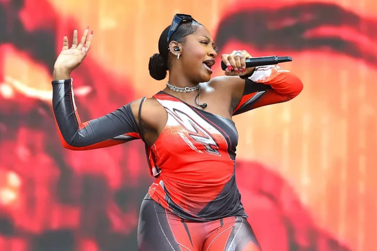 Una mujer glamurosa canta ante un micrófono. Lleva un conjunto rojo, negro y blanco ceñido al cuerpo y levanta una mano en el aire.