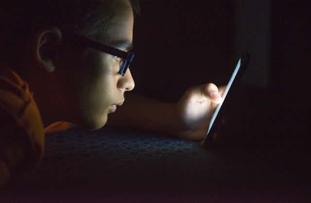 A teen boy is lying down as he looks at his cellphone in the dark.