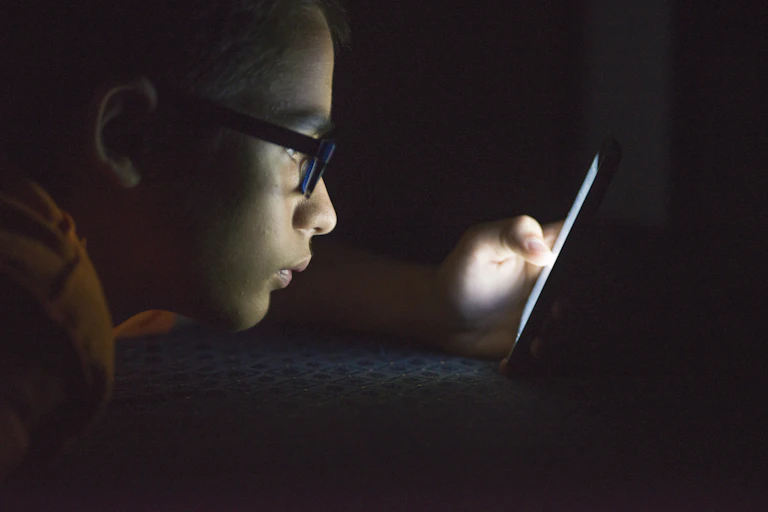 A teen boy is lying down as he looks at his cellphone in the dark.