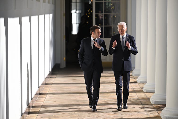 Two men in business suits, one with dark hair and one with white hair, gesture as they talk to each other and walk along a formal colonnade.