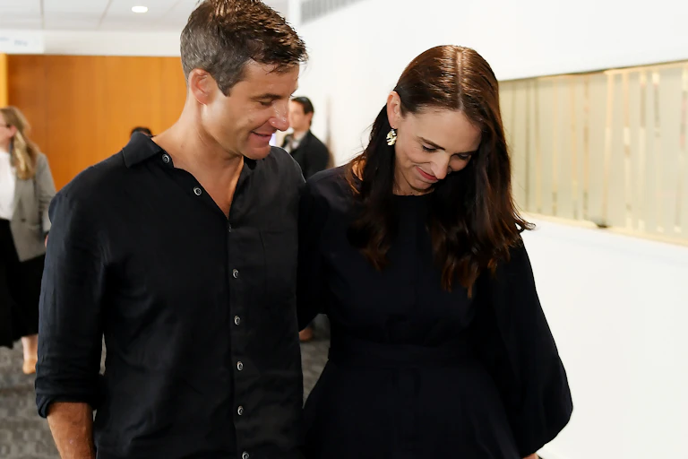 A white man and woman, both with dark hair and dark clothing, walk closely together.