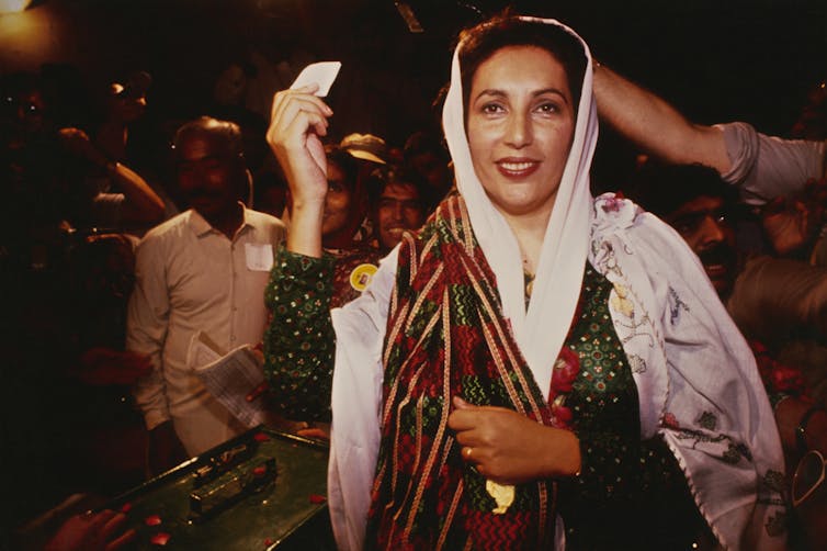 A woman with brown skin wears a headscarf and flowing clothing and holds up a small white piece of paper.