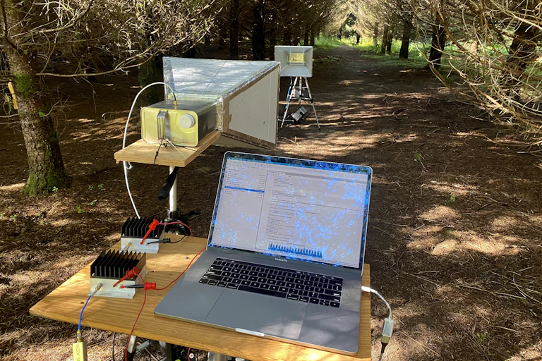 In a grove of trees, a laptops is open on a table in the foreground with a nearby rectilinear cone pointed toward the background where a similar rectilinear cone faces the foreground