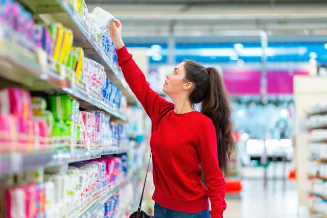 A woman in a red top reaches up for something on a shelf in a supermarket.