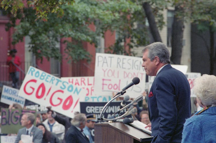 A man dressed in a suit speaks from a podium as people stand around holding banners.