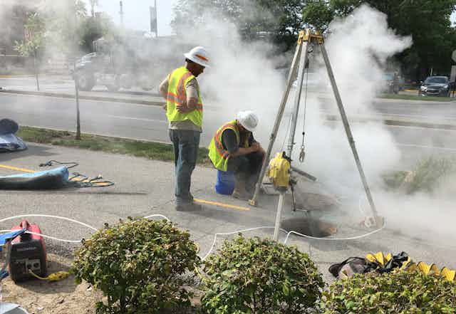 Two workers in construction vests work near a manhole with gases coming out. A liner and equipment are nearby.