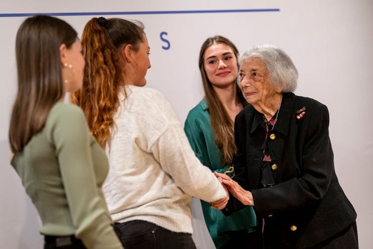 An elderly woman shakes hands with and chats with three teenage girls.