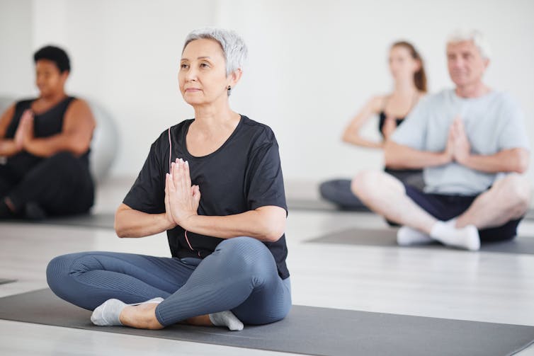 Un grupo de personas sentadas en colchonetas de yoga con las manos juntas frente al pecho.