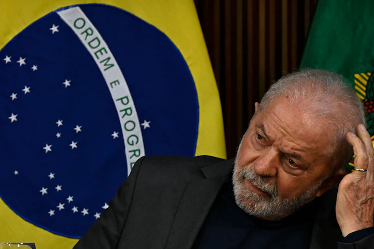 Man with gray hair and a beard looks worriedly to the side next to a flag that says 'ordem e progresso'