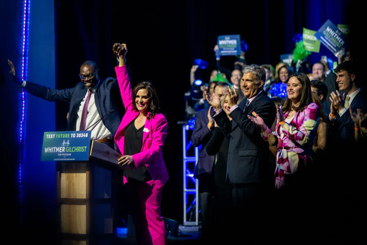 Standing behind a lectern, a smiling woman in a pink suit holds the raised hand of smiling, suited man