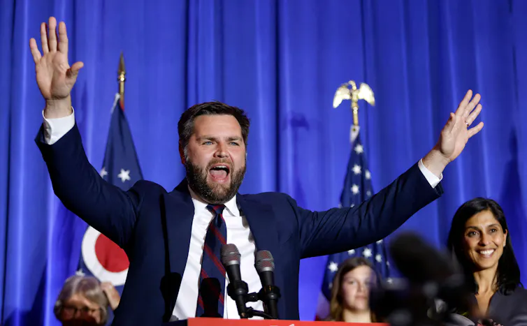 A suited man, with his mouth open, raises both hands, palms facing outward in celebration while a smiling woman and child stand behind him on his left
