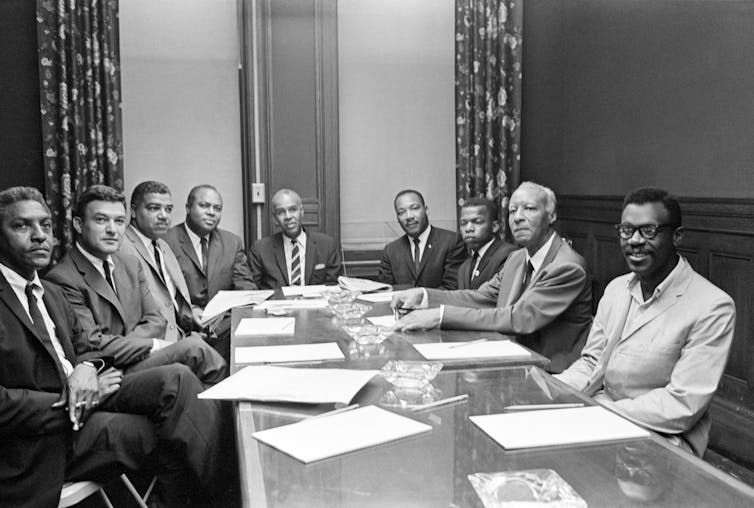 A group of men are sitting around a large table  with sheets of paper in front of them.
