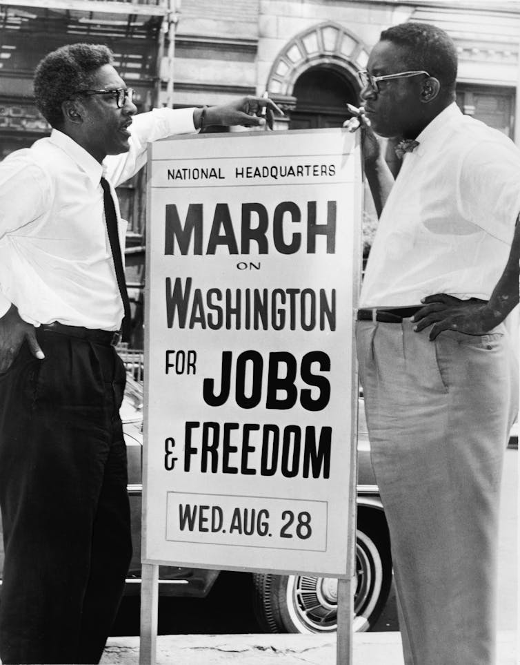 Two Black men are standing next to a sign that says March on Washington for jobs and freedom.
