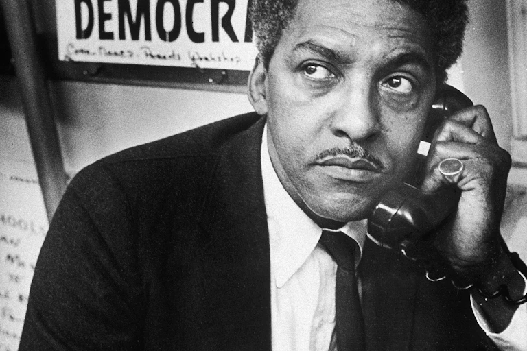 A black man wearing a dark jacket is talking on a telephone underneath a poster that says Jim Crow can't teach education.