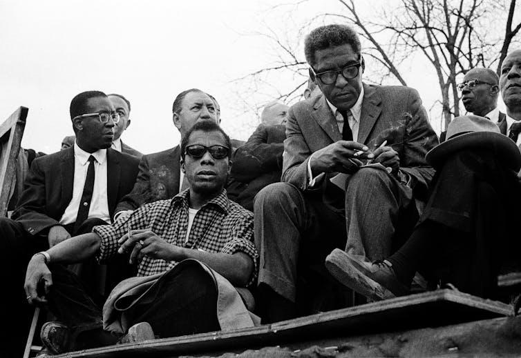 A Black man wearing sunglasses is sitting next to another Black man who is taking notes on a pad of paper.