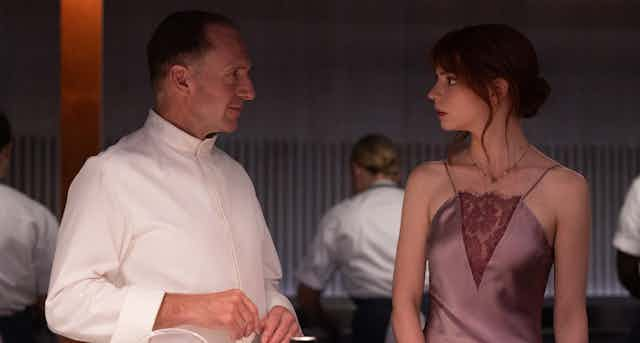 A man in chef whites stands next to a woman in a plum coloured halter satin dress. There are workers in the background and plating materials in the foreground.