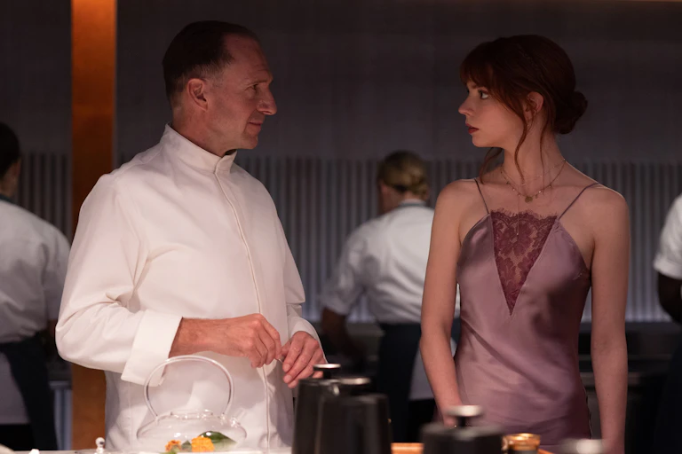 A man in chef whites stands next to a woman in a plum coloured halter satin dress. There are workers in the background and plating materials in the foreground.
